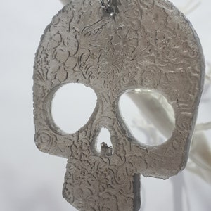 May include: A gray, skull-shaped ornament with floral detailing. The ornament has large eye sockets and a nose cavity. It hangs from a silver chain. The background is white.