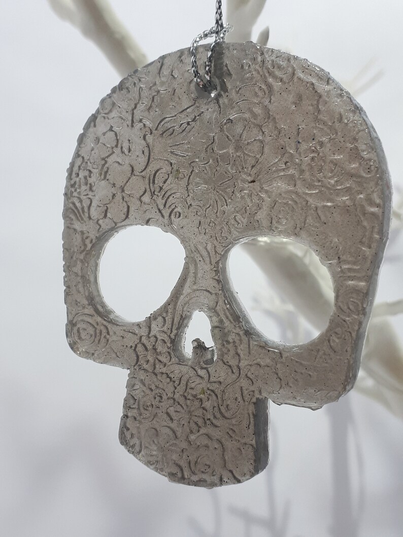 May include: A gray, skull-shaped ornament with floral detailing. The ornament has large eye sockets and a nose cavity. It hangs from a silver chain. The background is white.