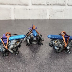 May include: Three small, gray, frog figurines with blue and purple accents. Each frog is wearing a backpack and carrying a small, blue, torpedo-shaped object.