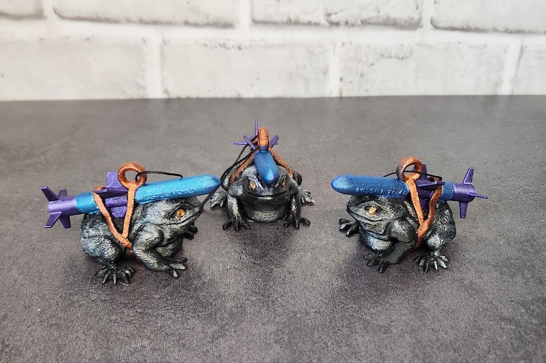 May include: Three small, gray, frog figurines with blue and purple accents. Each frog is wearing a backpack and carrying a small, blue, torpedo-shaped object.