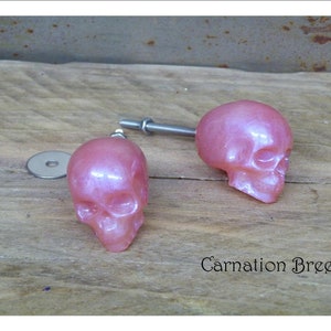 Peut inclure: Deux boutons de cabinet en forme de crâne rose avec un éclat métallique. Les boutons sont fixés à des vis argentées.