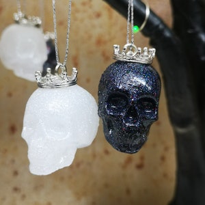 May include: Two glittery skull ornaments with silver crowns hanging from a branch. One skull is white and the other is black.