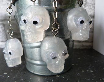 May include: Five clear plastic skull keychains with googly eyes hanging from a metal bucket. The skulls are translucent and have a white tint.