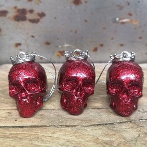 May include: Three red glitter skull ornaments with silver crowns. Each skull has a silver string attached for hanging.