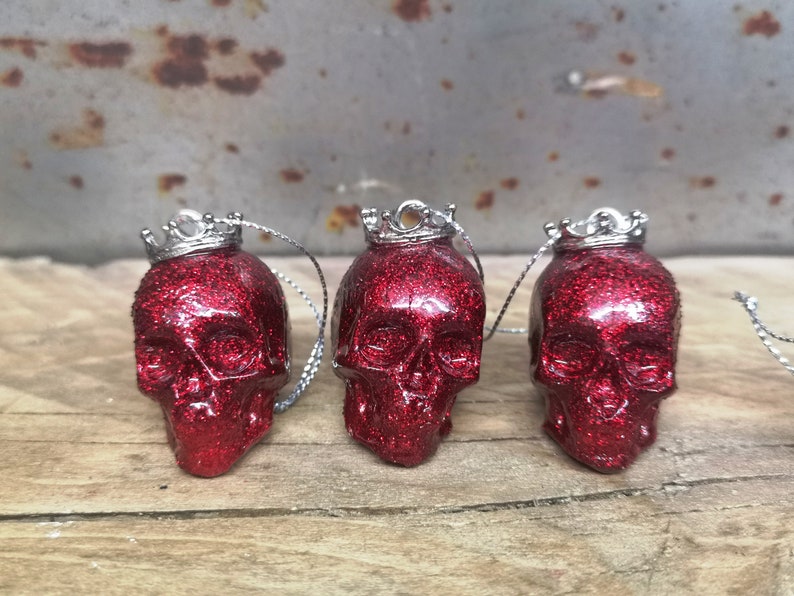 May include: Three red glitter skull ornaments with silver crowns. Each skull has a silver string attached for hanging.