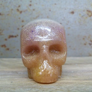 May include: A small, glittery, pink and gold skull made of resin. The skull has a detailed face with eye sockets, a nose, and a mouth.