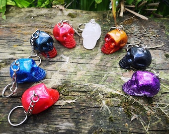 May include: A collection of skull-shaped keychains in various colors, including red, blue, black, and purple. Each keychain features a silver chain and ring. The keychains are displayed on a weathered wooden surface.