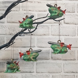 May include: Five green frog ornaments with red and silver missiles attached to their backs. The frogs are hanging from a black branch against a white brick wall.