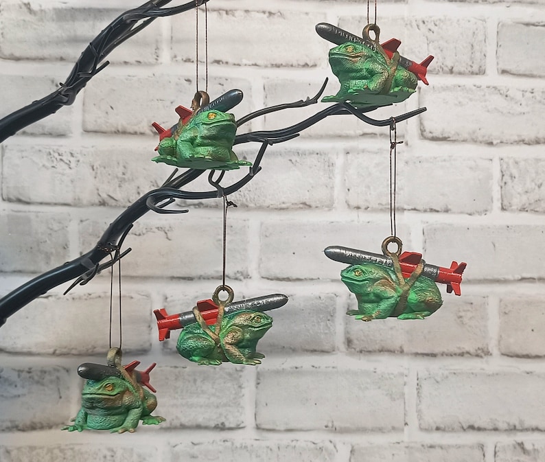 May include: Five green frog ornaments with red and silver missiles attached to their backs. The frogs are hanging from a black branch against a white brick wall.