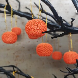 K&ouml;nnte beinhalten: Orangefarbene geh&auml;kelte Ornamente, die an schwarzen &Auml;sten h&auml;ngen. Die Ornamente sind rund und haben eine spitzenartige Textur.