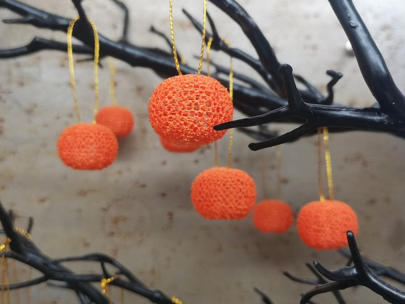 K&ouml;nnte beinhalten: Orangefarbene geh&auml;kelte Ornamente, die an schwarzen &Auml;sten h&auml;ngen. Die Ornamente sind rund und haben eine spitzenartige Textur.