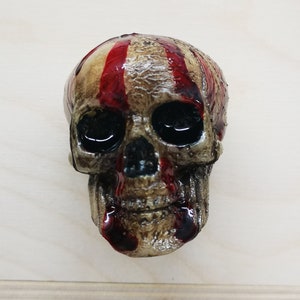 May include: A small, detailed skull figurine with red paint dripping down the face. The skull is made of a light brown material and has a textured surface.