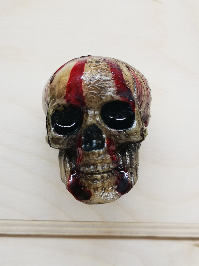 May include: A small, detailed skull figurine with red paint dripping down the face. The skull is made of a light brown material and has a textured surface.