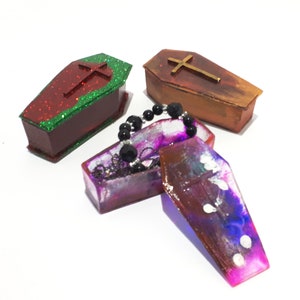 Pu&ograve; includere: Tre piccole scatole a forma di bara, una rossa con glitter verde, una marrone con glitter dorati e una viola con vortici bianchi e blu. Le scatole sono aperte e una collana di perle nere si trova all'interno di una delle scatole.