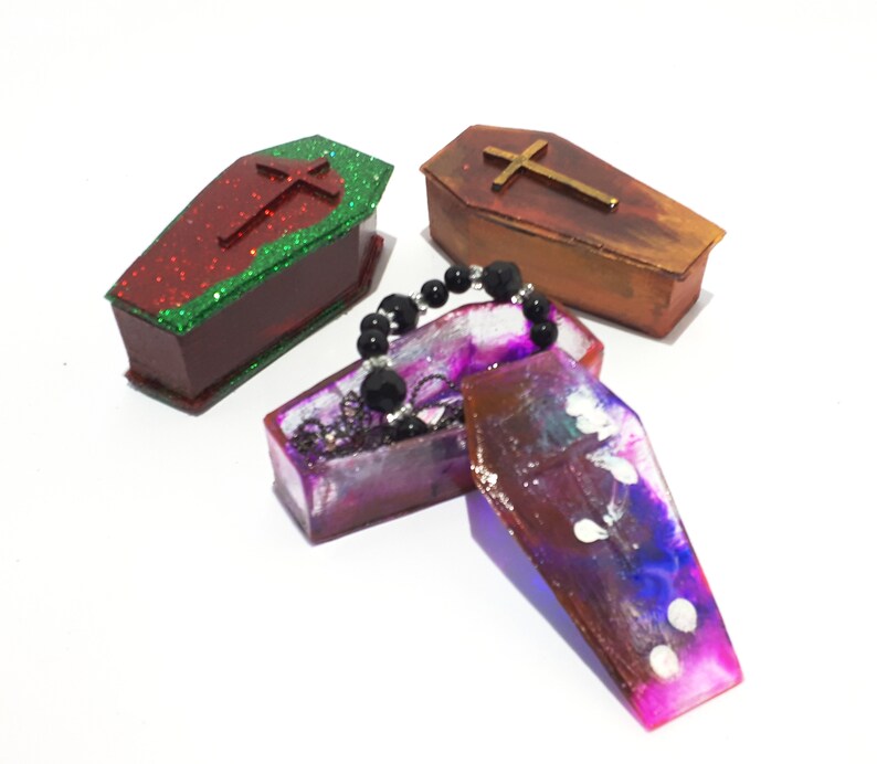 Pu&ograve; includere: Tre piccole scatole a forma di bara, una rossa con glitter verde, una marrone con glitter dorati e una viola con vortici bianchi e blu. Le scatole sono aperte e una collana di perle nere si trova all'interno di una delle scatole.