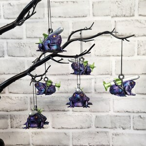 May include: Six purple frog ornaments with silver and green details hang from a black branch. Each frog is carrying a small silver missile.