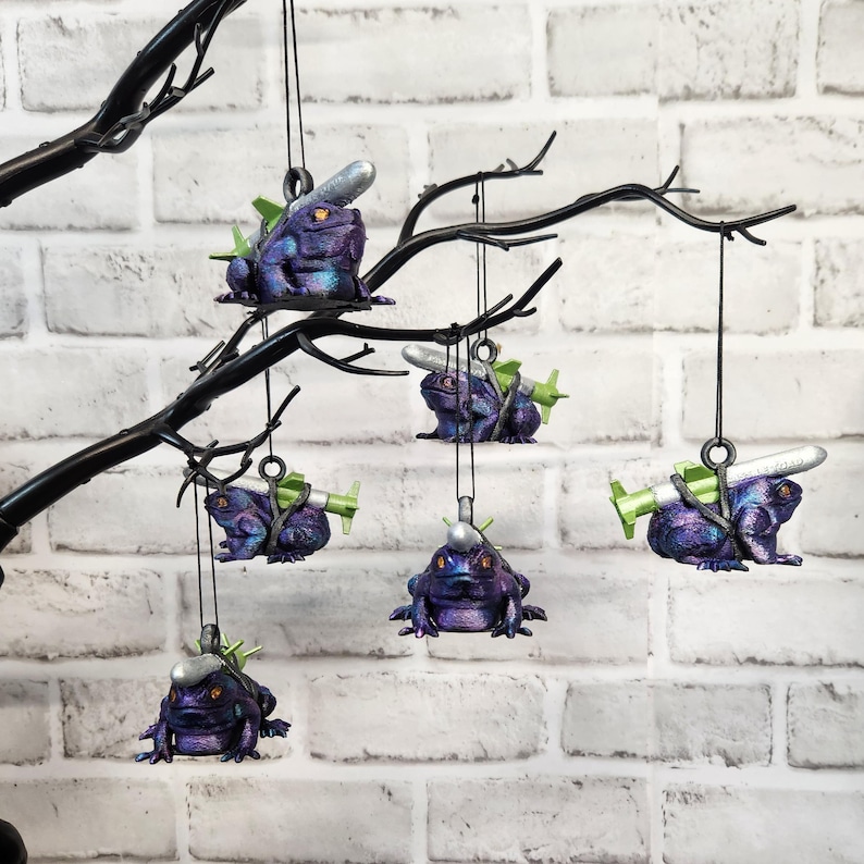 May include: Six purple frog ornaments with silver and green details hang from a black branch. Each frog is carrying a small silver missile.