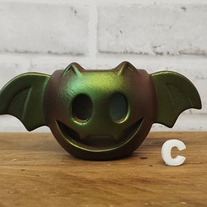 May include: A green and brown ceramic bat-shaped candle holder with a smiling face. The candle holder has a shimmery, iridescent finish.