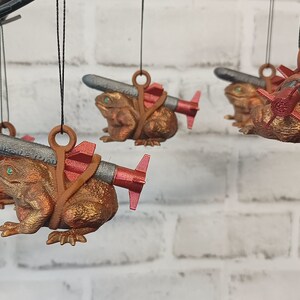 May include: Five brown frog ornaments with green eyes, each holding a small, silver-colored missile with red accents. The frogs are hanging from black strings.