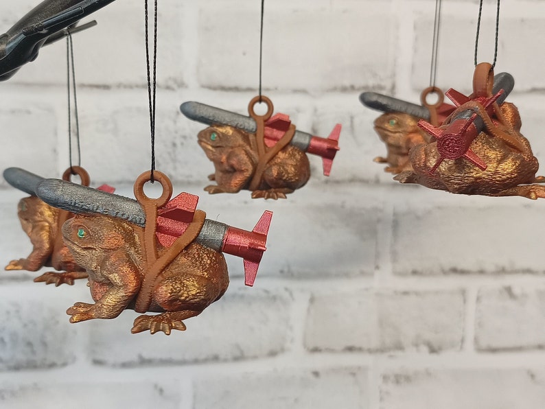 May include: Five brown frog ornaments with green eyes, each holding a small, silver-colored missile with red accents. The frogs are hanging from black strings.