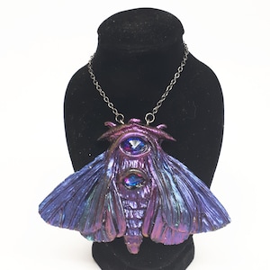 May include: A silver chain necklace with a large, iridescent purple and blue moth pendant featuring two large, faceted, blue gemstones for eyes.