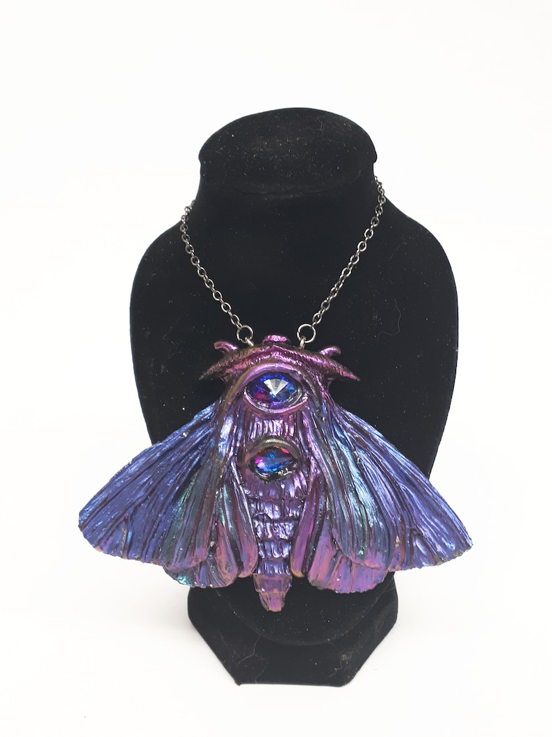 May include: A silver chain necklace with a large, iridescent purple and blue moth pendant featuring two large, faceted, blue gemstones for eyes.