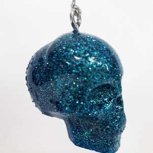 Puede incluir: Un adorno de calavera de color azul turquesa con purpurina y un lazo plateado para colgar.