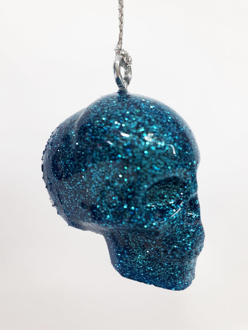 Puede incluir: Un adorno de calavera de color azul turquesa con purpurina y un lazo plateado para colgar.