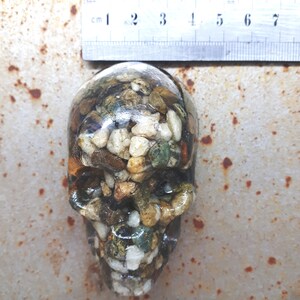 May include: A resin skull filled with small stones and pebbles in shades of brown, white, green, and gold. The skull is approximately 6 cm tall.