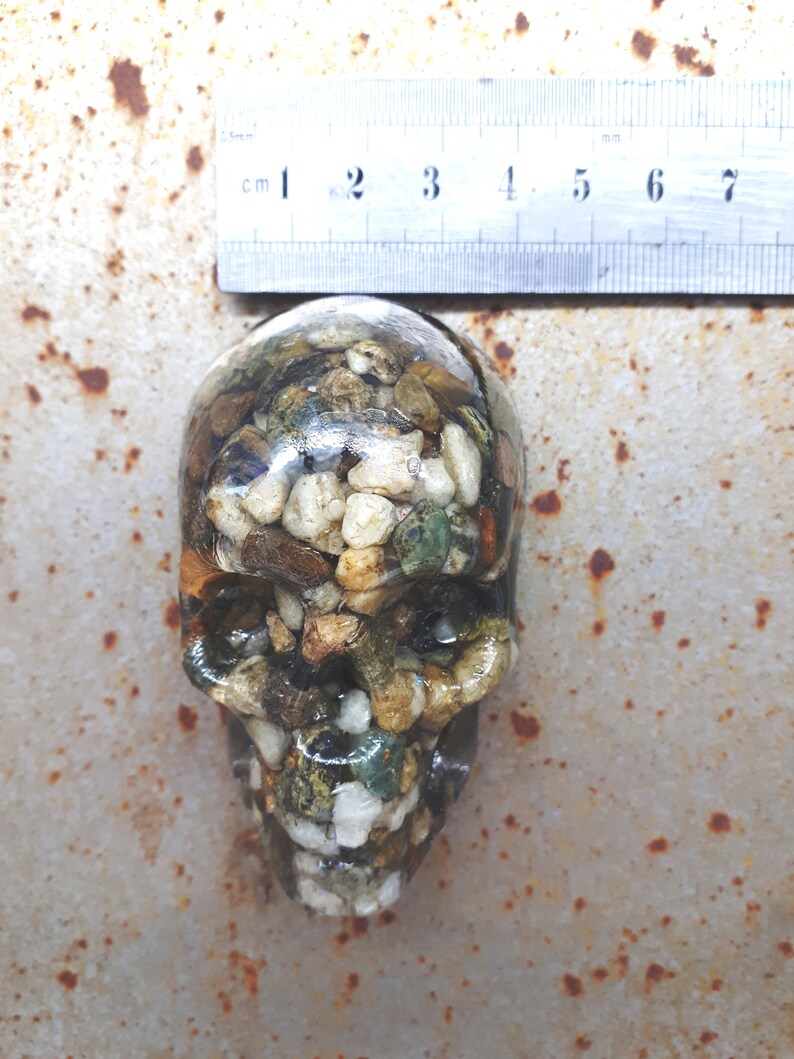 May include: A resin skull filled with small stones and pebbles in shades of brown, white, green, and gold. The skull is approximately 6 cm tall.