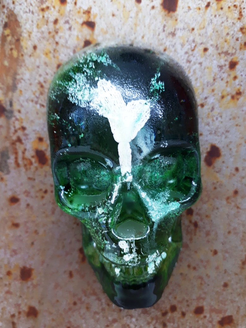 May include: A green glass skull with white paint accents. The skull has a detailed design and a textured surface.