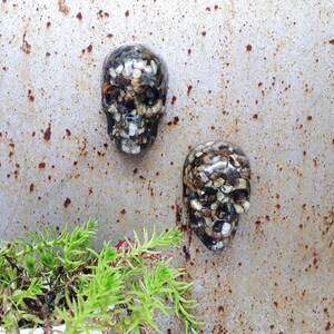 May include: Two resin skull ornaments filled with small stones. The skulls are black and white with a clear resin coating. The skulls are on a rusty metal background.