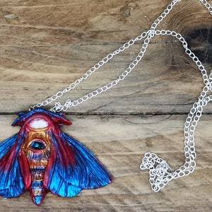 May include: A silver chain necklace with a blue and red moth pendant. The moth has a large, iridescent eye and intricate details.