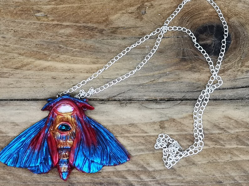 May include: A silver chain necklace with a blue and red moth pendant. The moth has a large, iridescent eye and intricate details.
