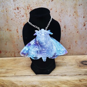 May include: A silver chain necklace with a large iridescent blue and purple moth pendant. The moth is made of resin and has a detailed, textured surface.
