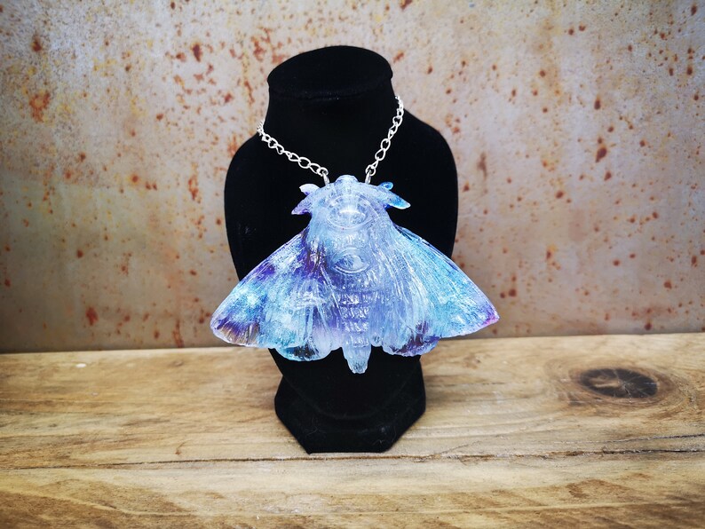 May include: A silver chain necklace with a large iridescent blue and purple moth pendant. The moth is made of resin and has a detailed, textured surface.