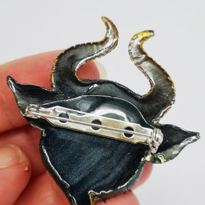 Puede incluir: Un broche de resina negro y verde con detalles dorados. El broche presenta una criatura cornuda estilizada con un broche de seguridad.