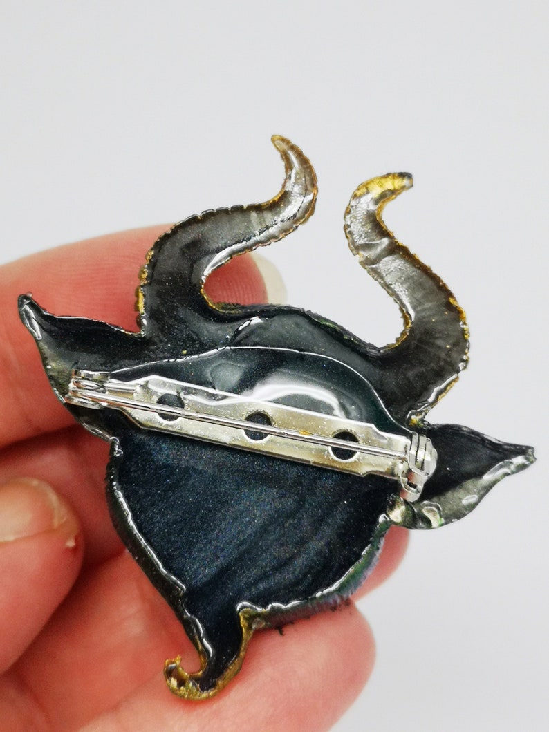 Puede incluir: Un broche de resina negro y verde con detalles dorados. El broche presenta una criatura cornuda estilizada con un broche de seguridad.