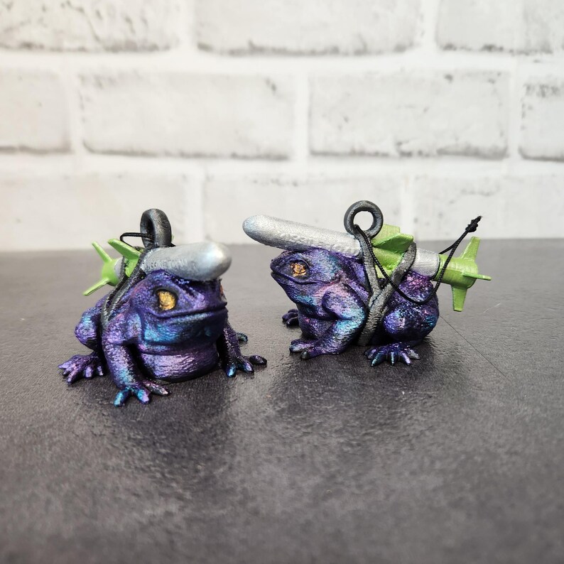May include: Two small, iridescent purple frog figurines with silver and green accents. Each frog is carrying a silver and green torpedo-shaped object on its back.