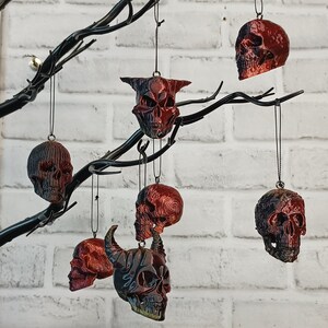 May include: A collection of six black and red skull ornaments hanging from a black metal branch. The skulls are all different shapes and sizes, and some have horns. The ornaments are hanging against a white brick wall.