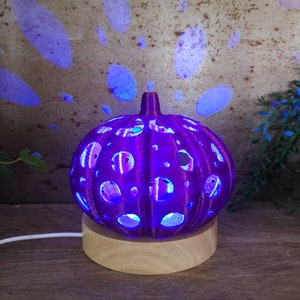 May include: A purple ceramic pumpkin-shaped lamp with a wood base. The lamp has a cut-out design and is lit from within, casting a blue glow.