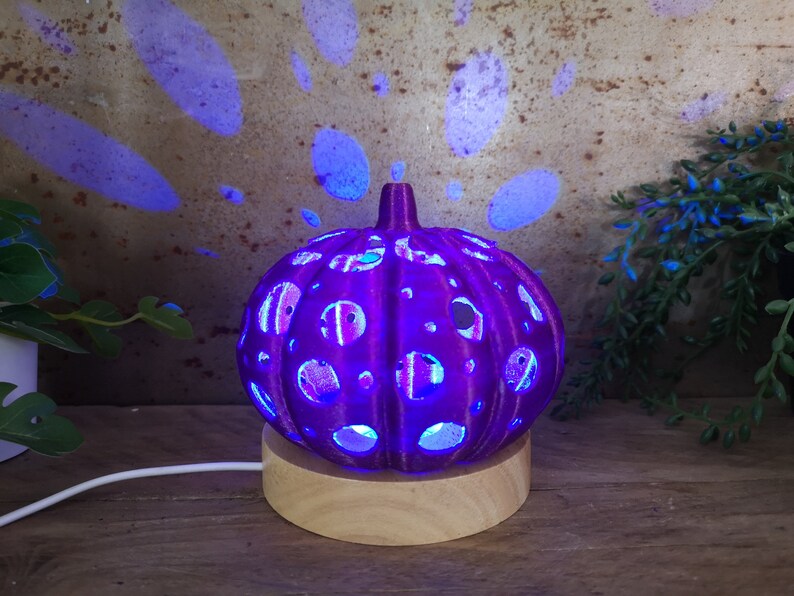May include: A purple ceramic pumpkin-shaped lamp with a wood base. The lamp has a cut-out design and is lit from within, casting a blue glow.
