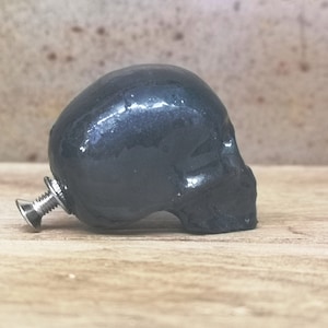 May include: A dark grey ceramic skull knob with a silver screw. The skull is glossy and has a detailed face.