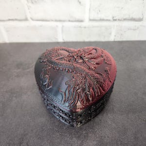 May include: A heart-shaped wooden box with a detailed carving of a dragon. The box is black with a red undertone and has a glossy finish.