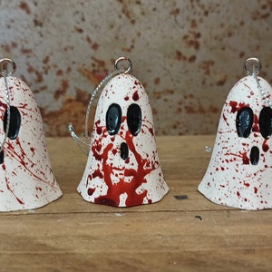 Puede incluir: Tres campanas blancas con forma de fantasma con ojos negros y pintura roja salpicada. Las campanas tienen cuerdas plateadas atadas a la parte superior.