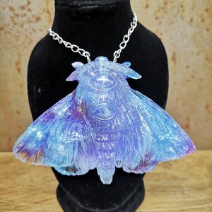 May include: A silver chain necklace with a blue and purple iridescent resin moth pendant. The moth has a detailed, textured design and a large, clear eye.