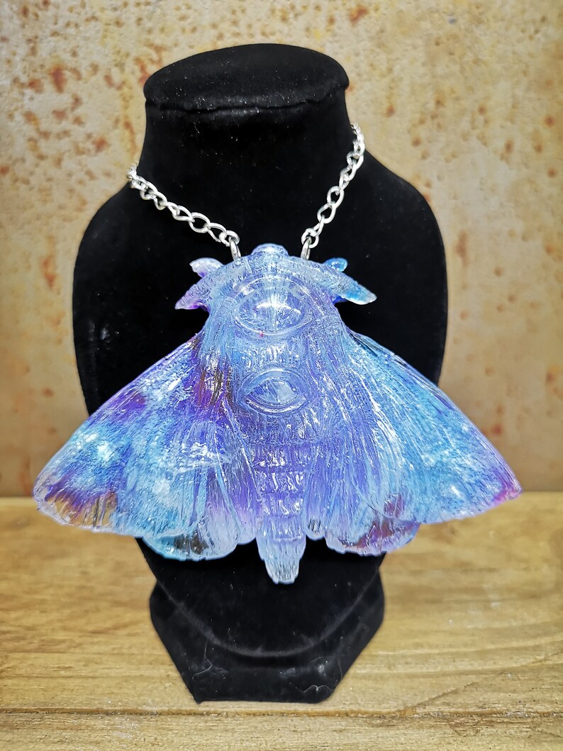 May include: A silver chain necklace with a blue and purple iridescent resin moth pendant. The moth has a detailed, textured design and a large, clear eye.