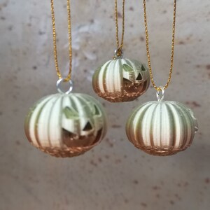 K&ouml;nnte beinhalten: Drei gr&uuml;n-wei&szlig; gestreifte K&uuml;rbis-Anh&auml;nger mit Jack-o'-Lantern-Gesichtern, die an goldenen Schn&uuml;ren h&auml;ngen.
