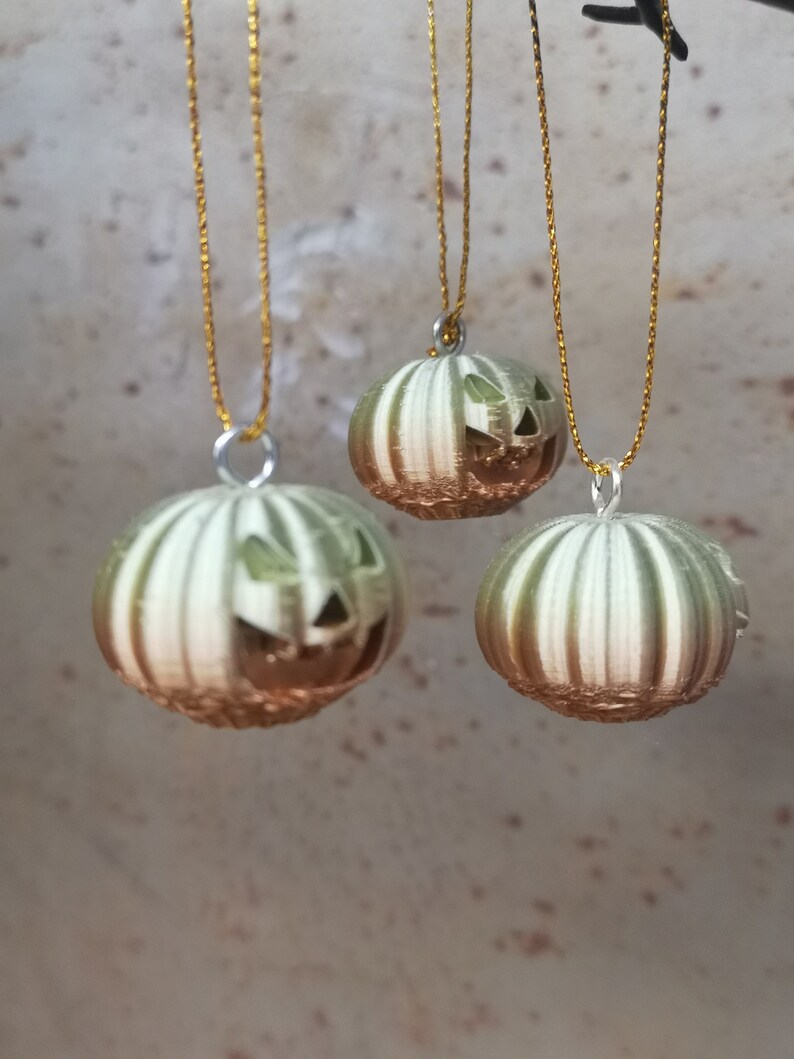 K&ouml;nnte beinhalten: Drei gr&uuml;n-wei&szlig; gestreifte K&uuml;rbis-Anh&auml;nger mit Jack-o'-Lantern-Gesichtern, die an goldenen Schn&uuml;ren h&auml;ngen.