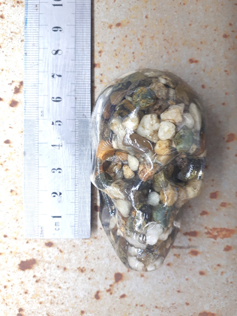 May include: A clear resin skull filled with small rocks and pebbles in shades of brown, white, and green. The skull is approximately 7 cm tall.